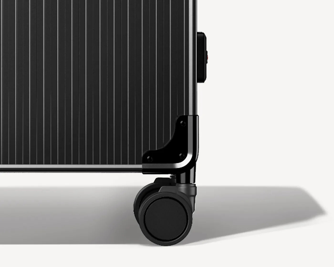Detail image of a black ribbed luggage lying on its side, focusing on the lower corner with a durable wheel and a black protective bumper, against a white background with a subtle shadow underneath indicating the suitcase is raised above the surface.